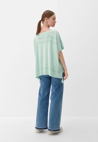 Licht mintgroene gebreide top met korte mouwen, voorzien van een gestructureerd patroon over de schouders en decoratieve franjes aan de onderrand.