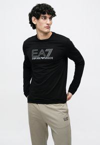 Must värviline pika varrukaga T-särk valge EA7 Emporio Armani logoga, mille juurde kuuluvad heledat beeži värvi püksid sama logoga. Lihtne, vabaaja disain.