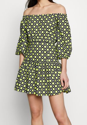 Femme portant une robe courte épaules dénudées avec des manches bouffantes trois-quarts, motif losanges noir et blanc avec des points jaune-vert.