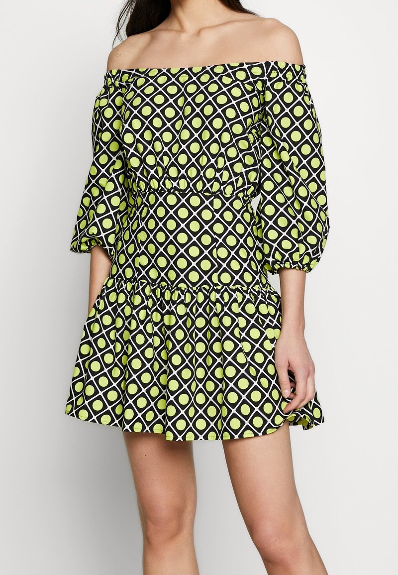 Femme portant une robe courte épaules dénudées avec des manches bouffantes trois-quarts, motif losanges noir et blanc avec des points jaune-vert.