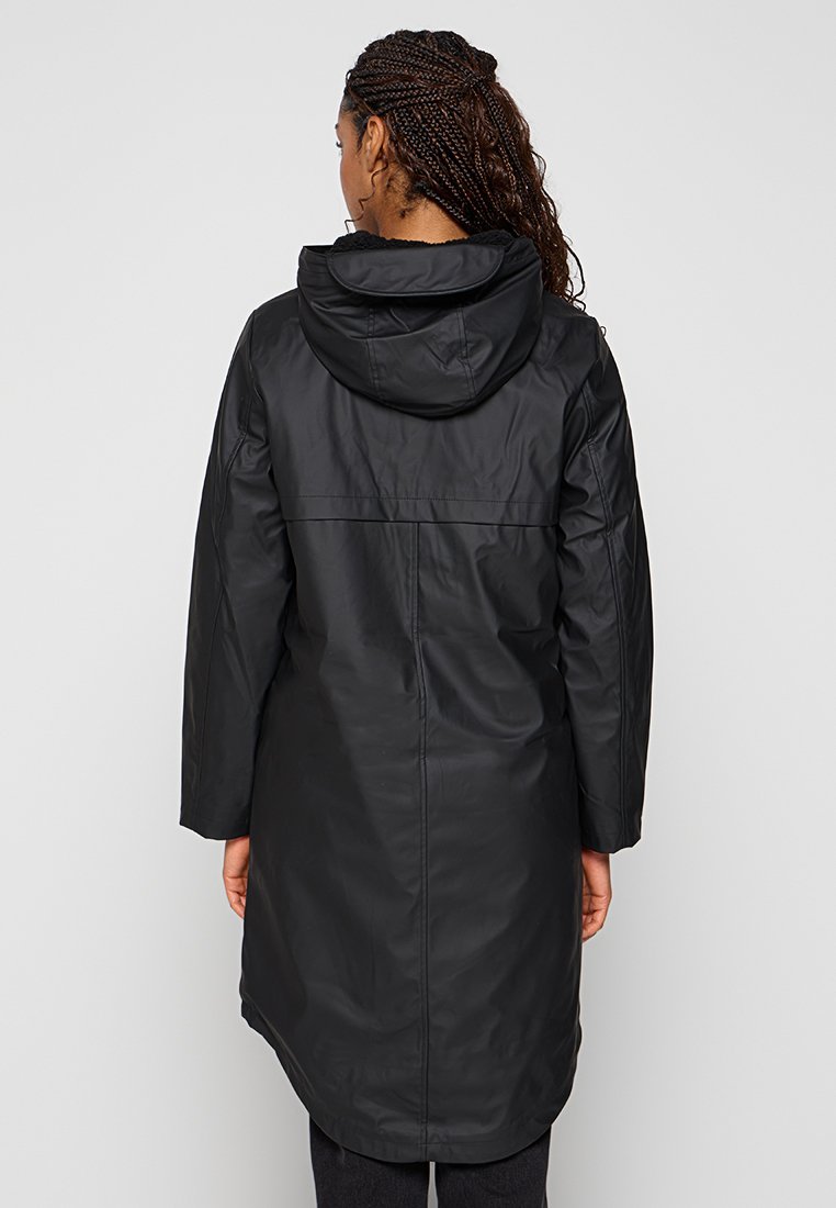 Femme aux cheveux tressés portant un long imperméable noir à capuche, se tenant de dos contre un fond clair et uni.