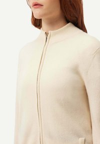 Krämfärgad zip-up-tröja i mjukt stickat tyg. Har hög krage och en minimalistisk design med en enkel fickdetalj.