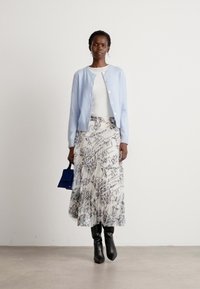 Lichtblauwe cardigan, witte top, gestreepte vloeiende rok in room en grijs, zwarte kniehoge laarzen, met een kleine blauwe handtas.