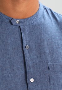 Camicia di lino blu con colletto alto, caratterizzata da una patta frontale con bottoni argentati e una tasca sul petto. Tessuto texturizzato con una trama sottile.