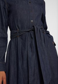 Jeansklänning i mörkblått med långa ärmar, knäppning framtill och ett självknutet bälte i midjan. Har kontrasterande sömmar och en avslappnad passform.