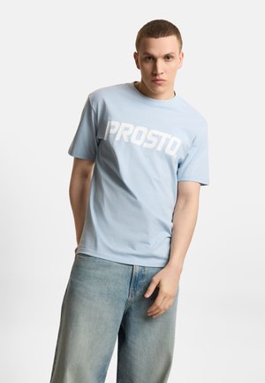 Młody mężczyzna w jasnoniebieskiej koszulce z logo "PROSTO" i luźnych, jasnych dżinsach, stojący pod kątem z jedną ręką wzdłuż ciała na jednolitym tle.