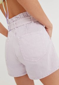 Femme portant un short en jean lilas taille haute avec de grandes poches arrière et une ceinture en tissu assortie sur fond blanc.