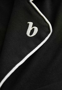 Fekete, puha tapintású szövet fehér pipás díszítéssel és a galléron fehér hímzett kisbetűs „b” betűvel.