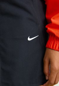 Juodi audiniai šortai su siuvinėta siūle, ant kurių yra balta Nike logo. Šone iš dalies matomas oranžinis ilgomis rankovėmis marškinėliai.