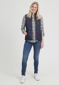 Gilet trapuntato blu navy con motivo a chevron, abbinato a una camicia a quadri grigia e jeans blu, completato da scarpe da ginnastica bianche.