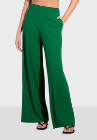 Zelene široke hlače izrađene od glatkog materijala, s visokim strukom i bočnim džepovima, uparene s crnim japankama.