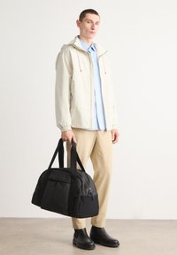 Svart duffelbag med doble håndtak og en glidelåslomme, med teksturert stoff. Modellen har på seg en lys beige jakke og beige bukser.