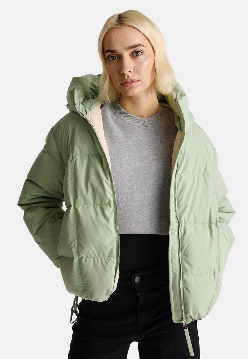 Jasnozielona kurtka puchowa z kapturem, z beżowym wykończeniem i zapięciem na zamek, noszona na szarym swetrze. Tkanina wydaje się błyszcząca i miękka.