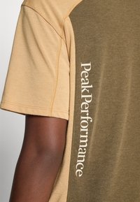 Delt kortermet skjorte med beige erm og olivengrønn kropp, med hvit vertikal "Peak Performance" tekst langs sidekanten.