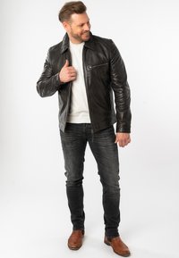 Zwarte leren jas met ritssluiting aan de voorkant, klein borstzakje en kraag. Draagt over een wit shirt, gecombineerd met donkere spijkerbroek en bruine laarzen.