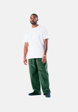 Uomo in piedi, con indosso una maglietta bianca, pantaloni verdi ampi, scarpe nere, orologio d'oro e un tatuaggio visibile sul braccio sinistro, su sfondo bianco.