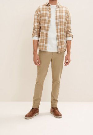 Bruin geruit overhemd over een lichte trui, gecombineerd met beige broek en bruine veterlaarzen. Eenvoudig ontwerp met neutrale kleuren en een gelaagde stof.