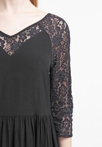Vestido negro con escote en V, que presenta mangas de tres cuartos hechas de encaje texturizado con patrones florales y una falda fluida.