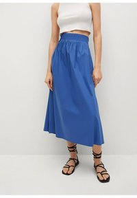 Donna che indossa un crop top bianco, una gonna midi blu a vita alta e sandali piatti neri con le bretelle, in piedi contro un muro chiaro.