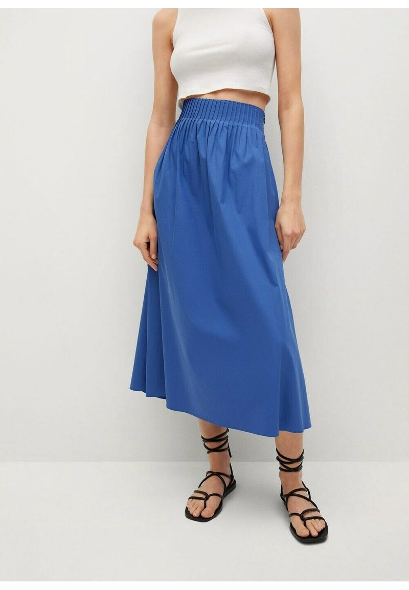 Donna che indossa un crop top bianco, una gonna midi blu a vita alta e sandali piatti neri con le bretelle, in piedi contro un muro chiaro.