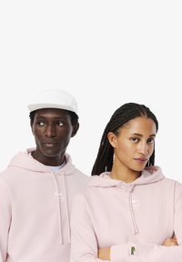 Sudaderas de color rosa claro en tejido suave, con un logo blanco de Lacoste y un emblema de cocodrilo verde en la muñeca. Una gorra blanca completa el conjunto.