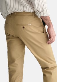 Beige bukser lavet af bomuld med en glat tekstur, der har to baglommer, hvoraf den ene har en brun knap og et logo-tag på linningen.
