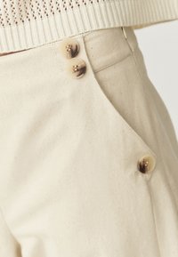 Pantalons en coton beige avec un design de poches pointues et trois boutons décoratifs à la taille. Texture lisse avec de légères speckles.