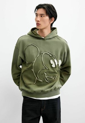 Junger Mann trägt olivgrünen Hoodie mit großem umrandetem Cartoon-Maus-Gesicht, schaut seitlich mit Händen hinter dem Rücken vor weißem Hintergrund.