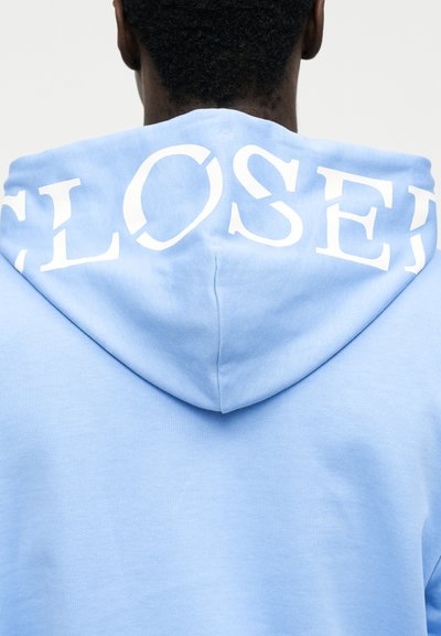 Personne vue de dos portant un sweat à capuche bleu clair avec le mot "CLOSED" imprimé en grandes lettres blanches sur la capuche.