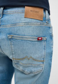 Jasnoniebieskie dżinsy denimowe z skórzaną naszywką Mustang na tylnej części pasa, z czerwonym emblematem nad tylną kieszenią oraz dekoracyjnym przeszyciem.