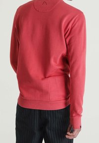 Sudadera roja de manga larga, con tejido texturizado, puños acanalados y cuello redondo. Incluye un pequeño bolsillo en la parte baja de la espalda.