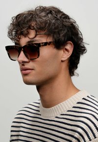 Occhiali da sole tartarugati con montatura quadrata e lenti sfumate, abbinati a un maglione a righe crema e navy. Capelli ricci visibili.