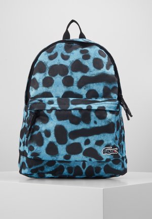 Sac à dos avec imprimé animal abstrait bleu et noir, une poche avant zippée, tirettes de fermeture éclair noires, et logo crocodile brodé en bas à droite.