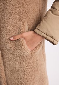 Karmelowy, puszysty płaszcz z owczej wełny z boczną kieszenią. Materiał ma teksturę, jest miękki i ma gładkie, beżowe rękawy.