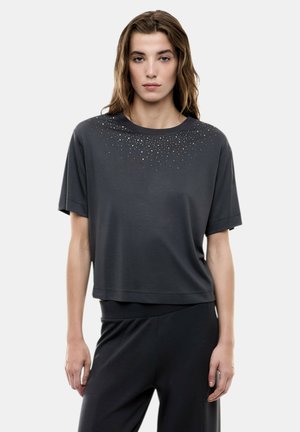 Donna con capelli lunghi che indossa una maglietta a maniche corte grigio scuro, ampia, decorata con piccoli borchiette lucide vicino allo scollo, e pantaloni scuri abbinati.