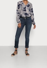 Donna che indossa una blusa a maniche lunghe navy a motivo paisley, jeans scuri arrotolati, cintura nera e sandali alla caviglia neri, in piedi su un pavimento grigio uniforme.