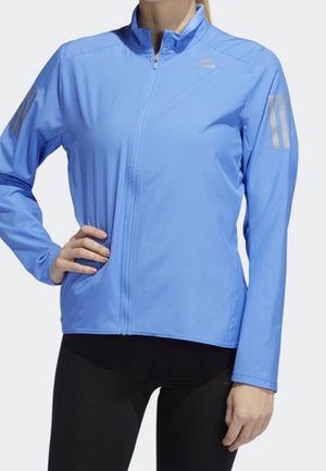 Veste de sport bleu clair à manches longues avec fermeture éclair, logo discret sur la poitrine et bandes réfléchissantes sur les deux manches, portée sur un legging noir.