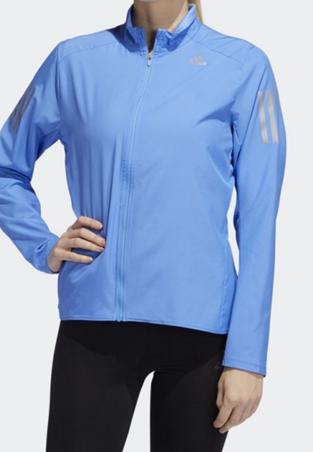 Veste de sport bleu clair à manches longues avec fermeture éclair, logo discret sur la poitrine et bandes réfléchissantes sur les deux manches, portée sur un legging noir.