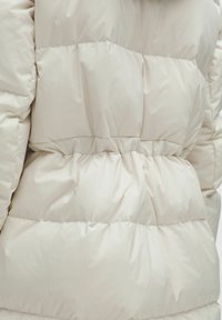 Bež puffer-jakk, millel on tikandiga disain, rõhutatud vöökoht ja pehme, läikiv materjal. Tekstuur näeb välja sile ja polsterdatud.