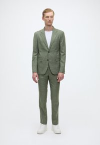 Hellgrüner Anzug aus strukturiertem Stoff, bestehend aus einem Zwei-Knopf-Blazer und passender Hose, kombiniert mit einem schlichten weißen T-Shirt und weißen Sneakern.