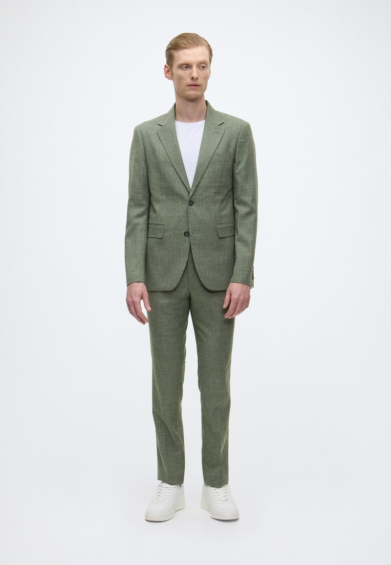 Hellgrüner Anzug aus strukturiertem Stoff, bestehend aus einem Zwei-Knopf-Blazer und passender Hose, kombiniert mit einem schlichten weißen T-Shirt und weißen Sneakern.