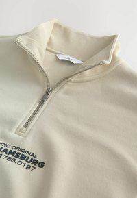 Jasno beżowa bluza rozpinana z prążkowanym kołnierzem, zapięcie na srebrny zamek i ciemnoniebieski nadruk z przodu. Miękki, teksturowany materiał.