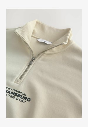 Svetlo bež zip-up jopica z ribastim ovratnikom, ki se ponaša s srebrnim zadrgo in temno modrim tiskanim besedilom na sprednji strani. Mehka, teksturirana tkanina.