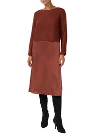 Langärmliges Kleid mit einem kurzen, strukturierten Oberteil in Rostfarbe und einem glatten, rostfarbenen Rock. Kombiniert mit knielangen Stiefeln aus schwarzem Wildleder.