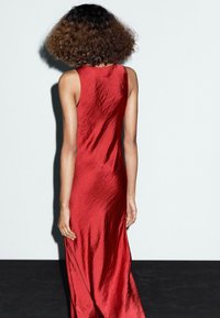 Une personne aux cheveux bouclés porte une longue robe rouge élégante, de dos, devant un fond uni avec un sol sombre.