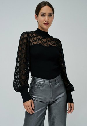 Top negro de manga larga con detalles de encaje y cuello alto, combinado con pantalones plateados de pierna ancha brillantes.