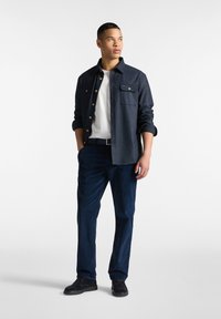 Marineblauwe overhem Dbno met twee borstzakken, witte t-shirt eronder, gecombineerd met donkerblauwe broek en zwarte schoenen, met een gladde textuur.