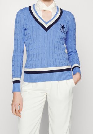 Person wearing a light blue cable-knit V-neck sweater with white and navy trim, white pants, and a white scarf tucked under the sweater.