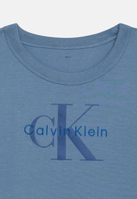 Kék pamut póló kerek nyakkal, amelyen kék hímzés olvasható: "Calvin Klein", valamint egy nagy, sötétebb árnyalatú "CK" nyomtatás található.