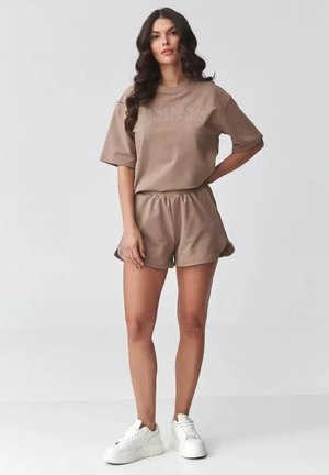 Kobieta stojąca z ręką na biodrze, ubrana w taupe-owy oversize'owy t-shirt i dopasowane szorty, białe sneakersy na platformie na jednolitym tle.